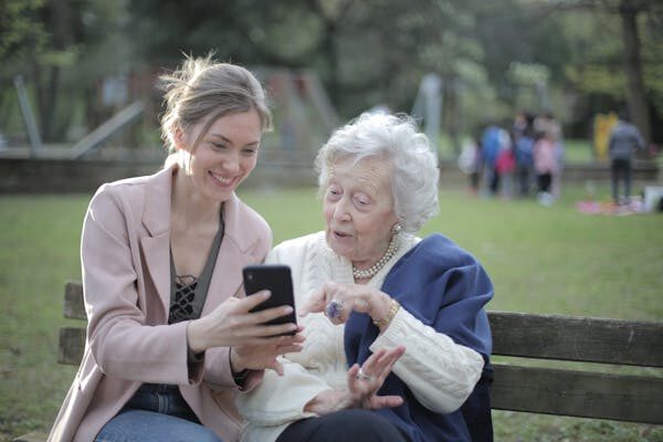 Junge und ältere Frau sitzen gemeinsam auf einer Bank im Park, junge Frau zeigt der älteren Frau lächelnd ihr Telefon